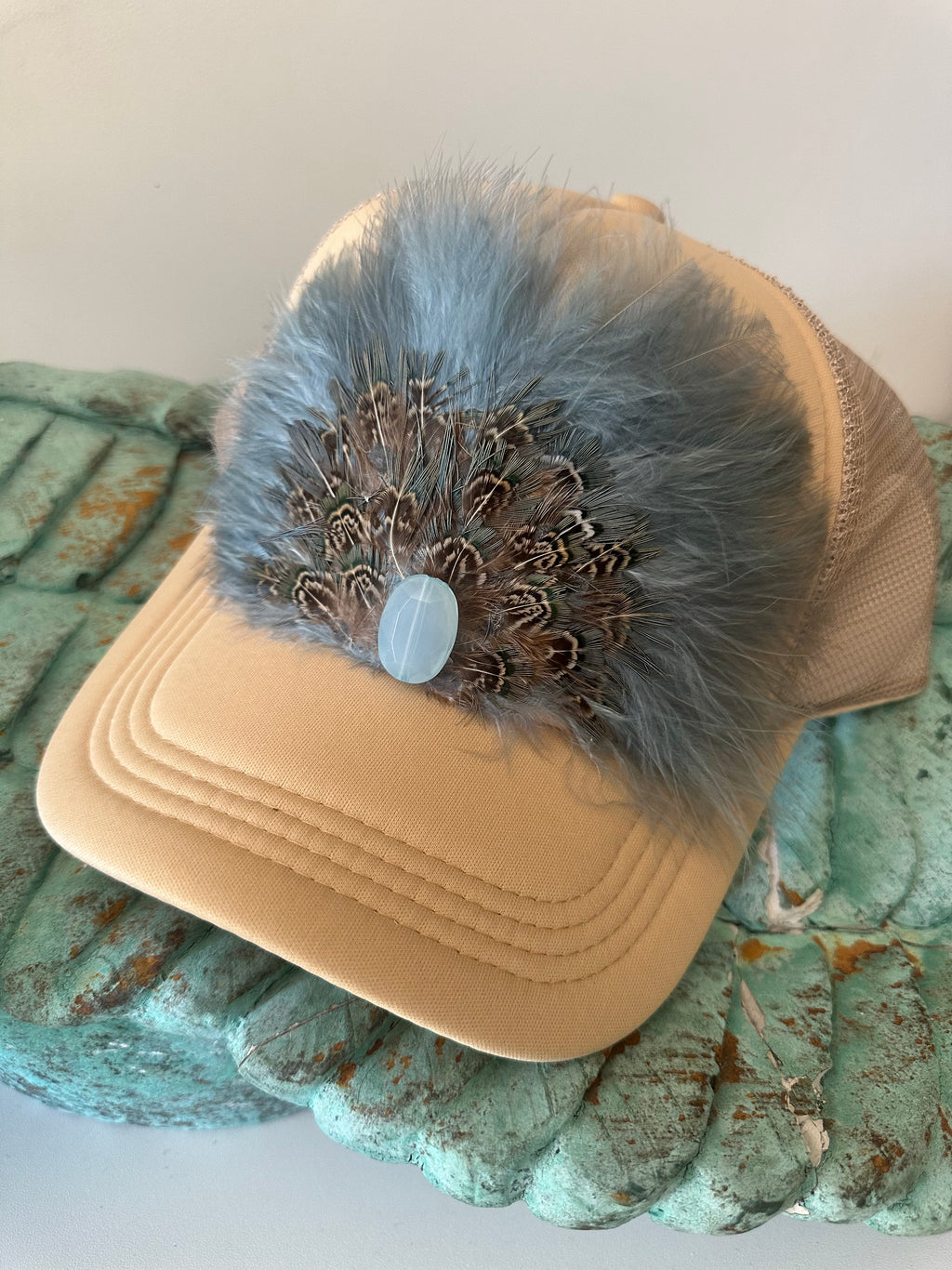 A beige trucker hat with dusty blue turkey feathers and blue and brown pheasant feathers. A light blue acrylic embelishment in the center. 